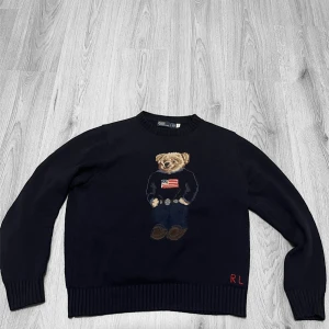Mörkblå stickad tröja Polo Bear RL - Mörkblå stickad tröja från Polo Ralph Lauren med ikoniska Polo Bear-motivet framtill. Tröjan har rund hals, ribbade muddar och RL-broderi nertill. Perfekt för dig som gillar klassisk preppy-stil med en twist.