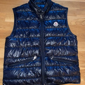 Mörkblå dunväst från Moncler - Snygg mörkblå dunväst från Moncler med glansig yta och quiltad design. Västen har två dragkedjefickor fram, hög krage och Moncler-logga på vänster bröst. Perfekt för lager-på-lager och har en stilren, sportig look. Färgen night blue och modellen gui. Storlek 2, uppskattas en medium av mig men skriv privat om du undrar över exakta mått. Säljer även på vinted om du hellre köper där