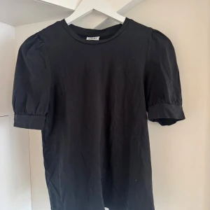 Svart topp med puffärm från Vero Moda - Säljer en svart topp från AWARE by Vero Moda i mjuk bomull. Modellen har puffiga korta ärmar med bred manschett och rund halsringning. Enkel och stilren design som funkar till många looks. Perfekt för dig som gillar minimalistisk stil.