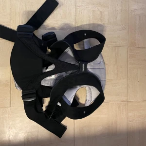 Babybjörn  - Bilderna visar en bärsele för barn, troligen från märket BabyBjörn. Produkten är svart med vita detaljer och har justerbara remmar för komfort och säkerhet. Passar barn mellan 3,5-4,5 kg (8-11 lbs). Selen ser ut att vara i gott skick utan synligt slitage eller defekter.
