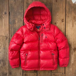 Röd pufferjacka från Polo Ralph Lauren - Säljer en fet röd pufferjacka från Polo Ralph Lauren med huva och dragkedja framtill. Jackan har två sidofickor med dragkedja och ikoniska Polo-loggan på bröstet. Perfekt för kalla dagar när du vill hålla stilen.