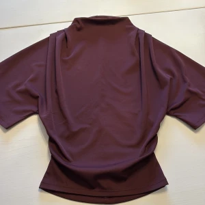 Vinröd polotröja med kort ärm - Snygg vinröd polotröja med korta ärmar och en hög krage. Tröjan har en stilren och enkel design med lätt drapering vid axlarna. Perfekt för dig som gillar minimalistiska plagg med en twist. Materialet känns mjukt och har ett lätt stretch.