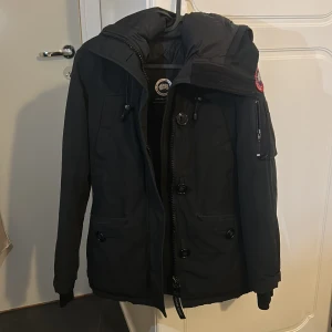 Svart dunjacka från Canada Goose - Svart dunjacka från Canada Goose med ikonisk patch på ärmen, dragkedja och knappar framtill, samt praktiska fickor och huva. Jackan har lång ärm och är perfekt för kalla dagar. Snygg och funktionell design som håller dig varm hela vintern. Tyvärr utan päls nypris 10 tusen  men 3000kr nu 