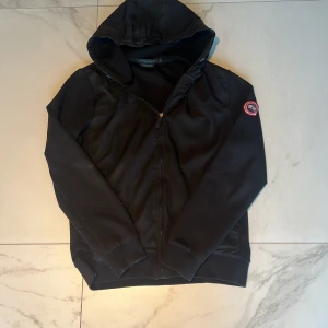 Svart vindjacka från Canada Goose - Svart Canada Goose Windbridge Hoody med huva och dragkedja framtill. Jackan har klassisk logga på ärmen och en clean, sportig look. Perfekt för dig som gillar stilrena och funktionella plagg. Snygg passform och ribbade muddar.