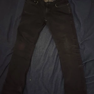 Svarta slim fit jeans från Nudie Jeans - Svarta jeans från Nudie Jeans med klassisk femficksdesign och smal passform. Jeansen har snygga kontrastsömmar och metallknapp med logga. Det finns en lagning i skrevet. Priset är inte hugget u sten.