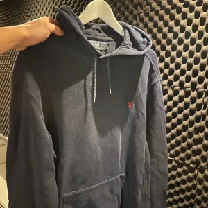 Mörkblå hoodie från Polo Ralph Lauren - Säljer en mörkblå hoodie från Polo Ralph Lauren med klassisk huva och snörning. Tröjan har en stor ficka framtill och den ikoniska röda Polo-loggan broderad på bröstet. Tillverkad i mjuk bomull, perfekt för chill dagar.