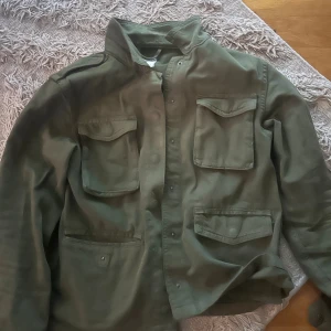 Grön overshirt med fickor - Säljer en grön overshirt med fyra stora fickor framtill och klassisk krage. Jackan har en loose passform och är tillverkad i bomull, perfekt för lager-på-lager. Stängs med knappar framtill och har långärmade ärmar.
