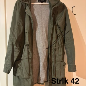 Grön parkajacka från bpc, strl 42 - Snygg grön parkajacka från bpc bonprix collection i storlek 42. Jackan har flera stora fickor, snörning i midjan och dragkedja framtill. Perfekt för dig som gillar en chill och praktisk stil. Materialet är bomull och jackan har långa ärmar.