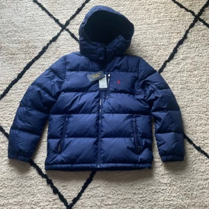 Mörkblå dunjacka från Polo Ralph Lauren - Snygg mörkblå dunjacka från Polo Ralph Lauren med huva och dragkedja framtill. Jackan har två sidofickor med dragkedja och det klassiska röda Polo-logot broderat på bröstet. Perfekt för kalla dagar och har en clean, puffad look.Aldrig använd då jackan var för liten vid inköp. 
