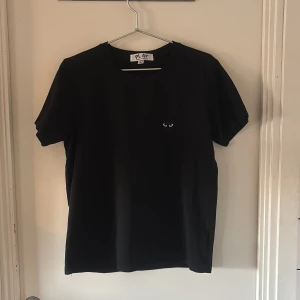 Svart Comme des Garçons Play t-shirt - Svart t-shirt från Comme des Garçons Play i storlek L men skulle säga att den sitter som ett M. Klassisk rund hals och korta ärmar. På bröstet finns det ikoniska broderade hjärtat med ögon i svart och vitt. T-shirten är gjord i mjuk bomull och har en clean, stilren look.