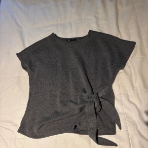 Grå stickad topp med knyt från Zara - Säljer en grå stickad topp från Zara i storlek S. Toppen har korta ärmar, rund halsringning och en snygg knytdetalj i sidan som ger en cool touch. Perfekt för dig som gillar enkel men trendig stil. Varan är lite små nopprig. 