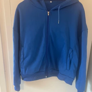 Blå hoodie med dragkedja och huva - Säljer en enkel blå hoodie med dragkedja och huva. Tröjan har långa ärmar, ribbade muddar och två fickor framtill. Perfekt för chill dagar och har en klassisk look med justerbar snörning i huvan.