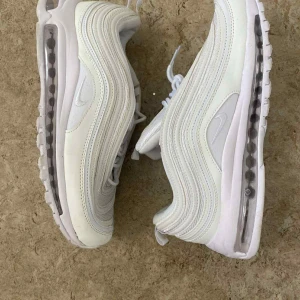 Nike Air Max 97 vita sneakers - Nike Air Max 97 sneakers i helvitt med ikoniska vågiga linjer och synlig Air-sula. Skorna har snörning, rund tå och platt sula. Materialmix av syntet och mesh ger en sportig vibe. Perfekta för dig som gillar clean och stilren look.