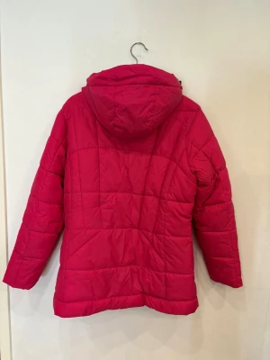Röd pufferjacka från On The Peak - Snygg röd pufferjacka med huva från On The Peak. Jackan har quiltad design, dragkedja framtill och praktiska fickor med dragkedja. Insidan är grå och huvan har justerbara snören. Perfekt för kalla dagar och riktigt chill stil.