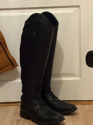Svarta ridstövlar i läder  - Säljer ett par svarta ridstövlar. Stövlarna har klassisk rund tå, snörning framtill och dragkedja baktill för enkel på- och avtagning. Snygg detalj med metallknapp med jh logga upptill. Perfekta för dig som gillar ridning.