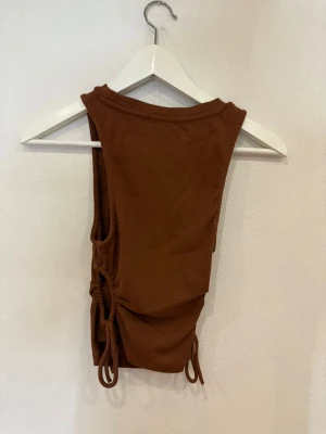 Brun ribbad linnetopp från Zara - Säljer en brun ribbad linnetopp från Zara med rund hals och ärmlös design. Toppen har snygga snörningsdetaljer på sidorna som går att justera för en cool look. Perfekt att styla med jeans eller kjol för en trendig vibe.