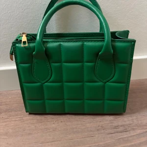 Grön quiltad handväska med dragkedja - Snygg grön handväska i fyrkantig modell med quiltad design och guldfärgad dragkedja. Väskan har två handtag och är tillverkad i syntetmaterial. Perfekt för dig som vill sticka ut med en färgglad accessoar.