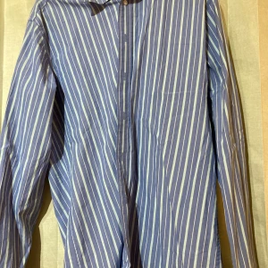 Blåvit randig skjorta från EASTWEST- NY - Säljer en blå skjorta med vita ränder från EASTWEST i storlek S. Skjortan har klassisk krage, långa ärmar och knäppning framtill. Perfekt för dig som gillar stilrena och fräscha plagg med lite edge.