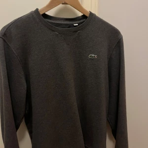 Mörkgrå sweatshirt från Lacoste Sport - Mörkgrå sweatshirt från Lacoste Sport med klassisk rund hals och det ikoniska gröna krokodilmärket broderat på bröstet. Tröjan har långärmad passform och är tillverkad i mjuk bomullsmix som är skön att bära. Perfekt för chill dagar och enkel att matcha. Storlek M och i väldigt bra skick🙌🙌