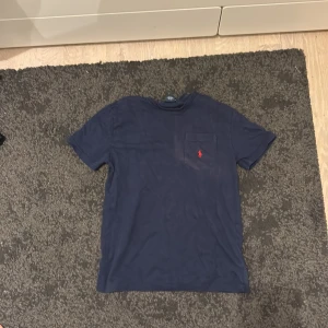 Marinblå t-shirt från Polo Ralph Lauren - Snygg marinblå t-shirt från Polo Ralph Lauren i 100% bomull. Klassisk passform med korta ärmar, rund hals och bröstficka med röd broderad logga. Perfekt för en clean och stilren look.