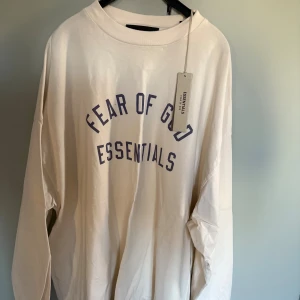 Beige långärmad tröja Fear of God - Säljer en beige långärmad tröja från Fear of God Essentials med tryck i mörkblått på bröstet. Tröjan har en loose passform och är tillverkad i mjuk bomull. Perfekt för dig som gillar streetwear och oversized stil.