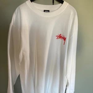 Vit långärmad Stüssy tröja - Vit långärmad tröja från Stüssy med röd logga på bröstet och stort tryck på ryggen. Tröjan har en klassisk rund hals och är gjord i mjuk bomull. Perfekt för dig som gillar streetwear och vill sticka ut med snygga detaljer.