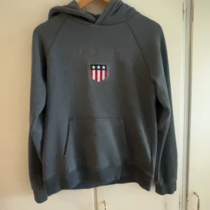 Grå hoodie från GANT med emblem - Snygg grå hoodie från GANT med broderat emblem på bröstet i rött, vitt och blått. Tröjan har huva, känguruficka och långa ärmar. Perfekt för chill dagar och har en klassisk, sportig look. Materialet är mjuk bomull med polyesterblandning.
