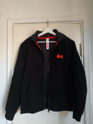 Sun 68 Bomber Double Hood - Svart hoodie från Sun 68 med orange tryck på bröstet och ärmen. Tröjan har huva med snörning, dragkedja framtill och en stor magficka. Perfekt för dig som gillar streetwear och vill ha en chill look. Tröjan är ny utan prislapp och i storleken large.Inköpt i Milano för 2200 kr. 