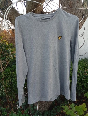 Grå långärmad tröja från Lyle & Scott - Snygg grå långärmad tröja från Lyle & Scott med klassisk rund hals och ikonisk gul broderad fågel på bröstet. Tillverkad i mjuk bomull, perfekt för lager-på-lager och chill vardagsstil.