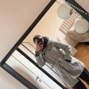 Grå kappa med huva från Zara Basic - Säljer en grå kappa från Zara Basic i storlek M. Den har en stor huva, knytskärp i midjan och dubbla knappar framtill. Kappan är långärmad och har en clean, stilren look som är perfekt för höst och vinter. Materialet är mjukt och känns som ullblandning.