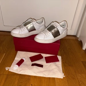 Valentino Open - Snygga Valentino open  i vitt skinn med bred silverfärgad detalj över vristen och klassiska nitar på hälen. Platt sula och rund tå, med snörning framtill. Alla Og saker följer med vid snabb och smidig affär 