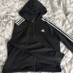 Svart Adidas hoodie med dragkedja - Svart hoodie från Adidas med klassiska vita ränder längs ärmarna och Adidas-logga på bröstet. Tröjan har huva med snörning, hel dragkedja och långa ärmar. Materialet är mjuk polyester, perfekt för chill eller träning.