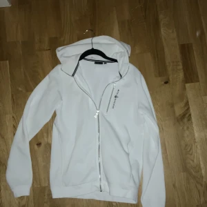 Vit hoodie från Sail Racing med dragkedja - Snygg vit hoodie från Sail Racing med hel dragkedja framtill och broderad logga på bröstet. Tröjan har huva, långa ärmar och mjukt fleecefoder på insidan. Perfekt för dig som gillar sportig och clean stil.