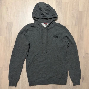 Mörkgrå hoodie från The North Face - Säljer en snygg mörkgrå hoodie från The North Face med klassisk logga på bröstet. Tröjan har huva med snörning, stor magficka och långa ärmar med ribbade muddar. Mjuk insida i bomullsmix som gör den extra skön. 