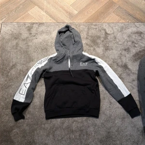 EA7 hoodie med half zip och logga - Snygg hoodie från EA7 med half zip och huva. Färgblockad i svart, grått och vitt med stora loggor på ärmen och bröstet. Långärmad och med känguruficka framtill. Perfekt för dig som gillar streetwear och sportig stil.
