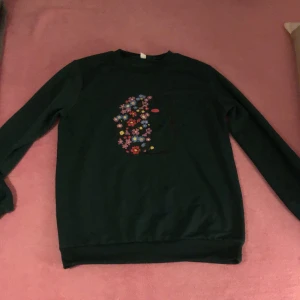 Mörkgrön sweatshirt med blommigt igelkott - Mörkgrön sweatshirt med lång ärm och rund hals. På framsidan finns ett stort tryck av en igelkott fylld med färgglada blommor i rosa, rött, blått och gult. Tröjan har en klassisk passform och är gjord i mjuk bomull, perfekt för dig som älskar att styla dina kläder och hitta din stil 