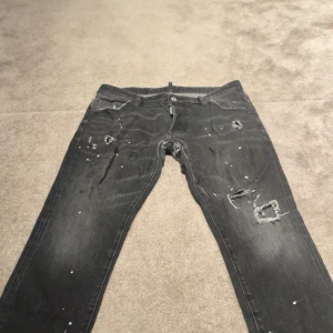 Svarta jeans från Dsquared2 med slitningar - Svarta jeans från Dsquared2 med coola slitningar och hål på både fram- och baksida. Jeansen har vita färgstänk för en edgy vibe och klassisk femficksdesign. Passar dig som gillar streetstyle och vill sticka ut med detaljerad look. Storlek S/M