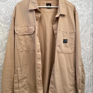Beige overshirt från Lee Workwear - Snygg beige overshirt från Lee Workwear med diskret fiskbensmönster. Skjortan har två bröstfickor med knappar och Lee-logga, klassisk krage och knäppning framtill. Tillverkad i mjuk bomull som känns skön mot huden. Perfekt att slänga på över en t-shirt.