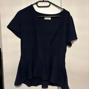 Mörkblå omlottblus med volang - Snygg mörkblå omlottblus från Object Collectors Item med korta ärmar och volang nertill. Blusen har v-ringning och en lätt struktur i tyget som ger en stilren look. Perfekt att styla med jeans eller kjol för en trendig outfit.