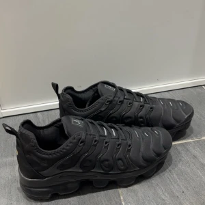 Nike Vapormax svarta sneakers - Svarta Nike Vapormax sneakers med chunky, genomskinlig sula och coolt mönstrad ovandel. Skorna har snörning och detaljer i syntetmaterial, samt en rund tå. Perfekt för dig som gillar sportig och modern stil. Diskret Nike-logga på sulan och VM-märke bak.