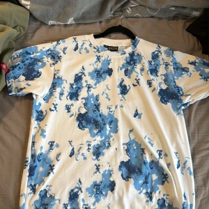 Vit och blå t-shirt från Excite - Säljer en vit t-shirt från Excite med blått camouflagemönster och tryckta bokstäver över hela plagget. T-shirten har rund hals och korta ärmar, perfekt för dig som gillar streetwear vibes. Materialet är mjuk bomull för skön känsla hela dagen.