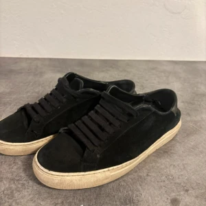 Svarta snickers från Axel arigato  - Svarta sneakers med ovandel i mjuk mocka och klassisk snörning. Skorna har en rund tå och vit platt sula som ger en cool kontrast mot det svarta. Perfekt för dig som gillar stilrena och enkla sneakers med en urban vibe.