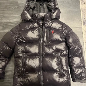 Svart pufferjacka från Polo Ralph Lauren - Svart pufferjacka från Polo Ralph Lauren med glansig finish, huva och flera dragkedjeförsedda fickor. Jackan har det klassiska röda Polo-logot broderat på bröstet och är fodrad för extra värme. Perfekt för kalla dagar med stil.
