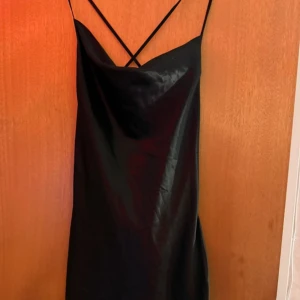 Svart satin slipklänning H&M Divided - Svart slipklänning i satin från H&M Divided med tunna axelband som korsas i ryggen. Klänningen har en snygg draperad urringning och är kort i modellen. Perfekt för dig som gillar minimalistisk och trendig stil.