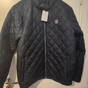 Mörkblå quiltad jacka från Vinson - Quiltad jacka från Vinson i mörkblå färg, storlek XL. Jackan har ett snyggt rutmönster, dragkedja framtill och två sidofickor. Märkespatch med Vinson-logga på bröstet. Perfekt för dig som gillar stilren och sportig look.