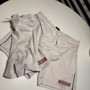  Två Vita sportshorts Team Sportia M - Säljer två par vita sportshorts från Team Sportia i storlek M. De har snörning i midjan, svart piping längs sidorna och är gjorda i ett luftigt meshmaterial som är perfekt för träning. Team Sportia-logga finns broderad på benet.