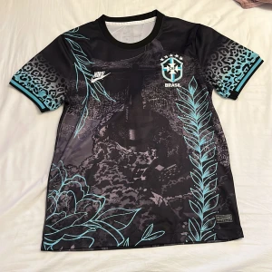Nike Brasil fotbollströja svart/blå - Snygg Brasil fotbollströja från Nike i svart med turkosa detaljer och leopardmönster på ärmarna. Tröjan har korta ärmar, rund hals och coolt grafiskt tryck med Kristusstatyn och blommor. Materialet är lätt och andas, perfekt för match eller träning.