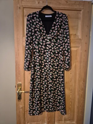 Blommig midiklänning från Adoore - Supersnygg långklänning från Adoore med svart botten och rosa blommor. Klänningen har v-ringning, långa ärmar och knappar framtill. Den har en feminin siluett och är perfekt för dig som gillar romantiska detaljer och blommigt mönster.