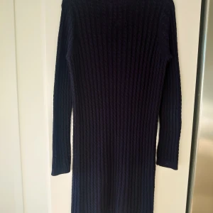 Mörkblå stickad klänning Lexington S - Supersnygg mörkblå stickad klänning från Lexington i storlek S. Klänningen har långärmade ärmar och ett ribbat kabelstickat mönster som ger en chill och klassisk vibe. Perfekt för dig som gillar enkel men stilren design med diskret Lexington-logga i metall på ärmen.