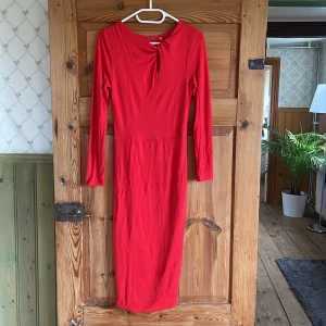Röd långärmad klänning med twist - Snygg röd långklänning med lång ärm och twistad detalj framtill vid halsen som skapar en liten öppning. Klänningen har en figurnära passform och stängs med dragkedja i ryggen. Materialet är mjukt och stretchigt, troligen polyester. Perfekt för dig som vill sticka ut.